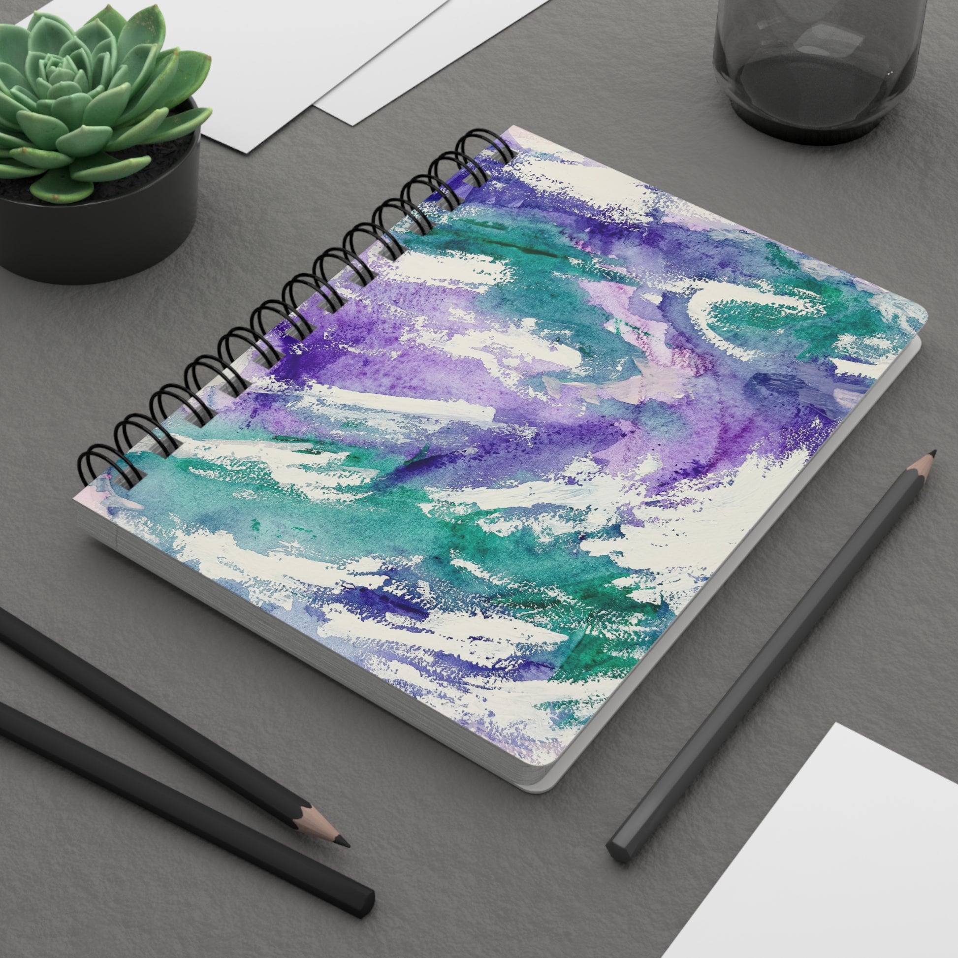 A spiral-bound notebook with a abstract painting cover featuring flowing textures of deep purple, emerald greens, and white. On a desk.