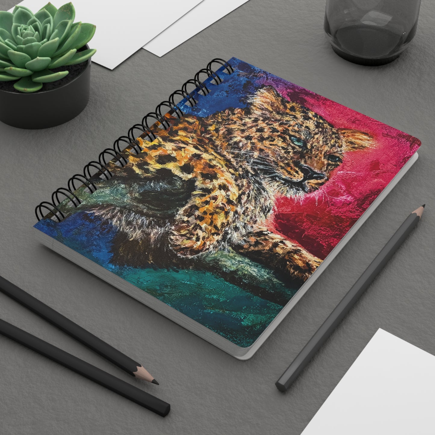 A spiral notebook with a painting of a jaguar with rich pinks greens and blues. On a desk.