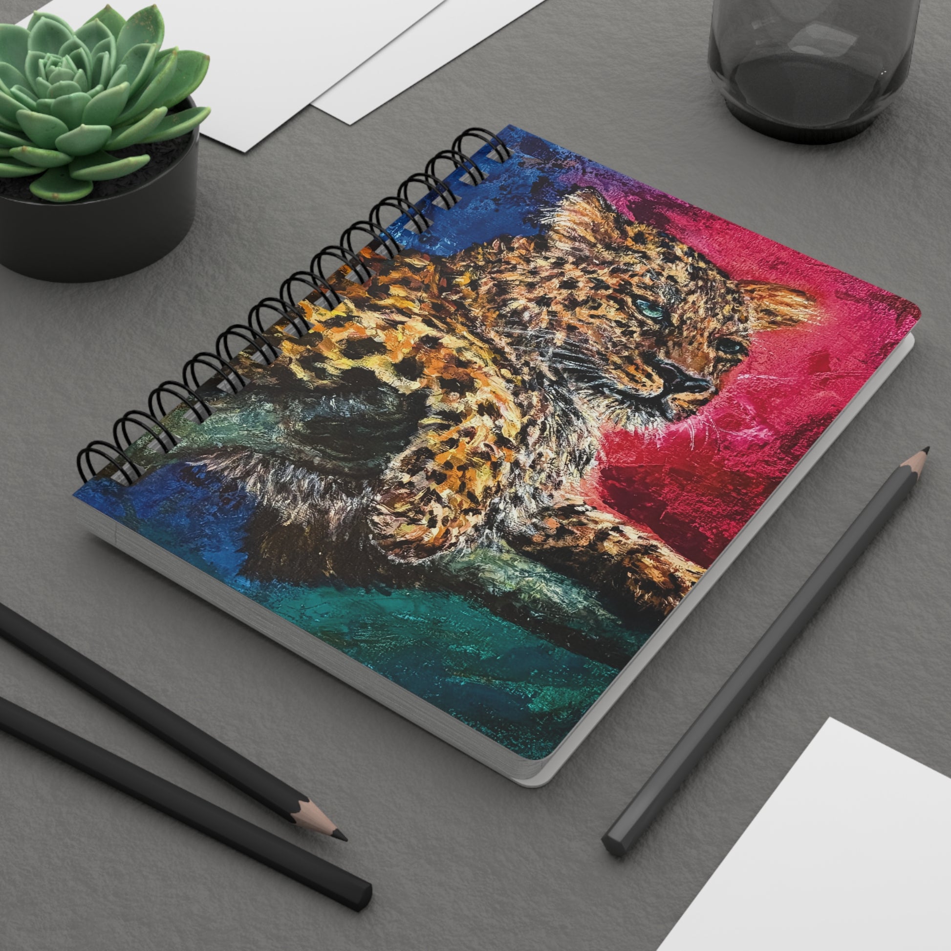A spiral notebook with a painting of a jaguar with rich pinks greens and blues. On a desk.