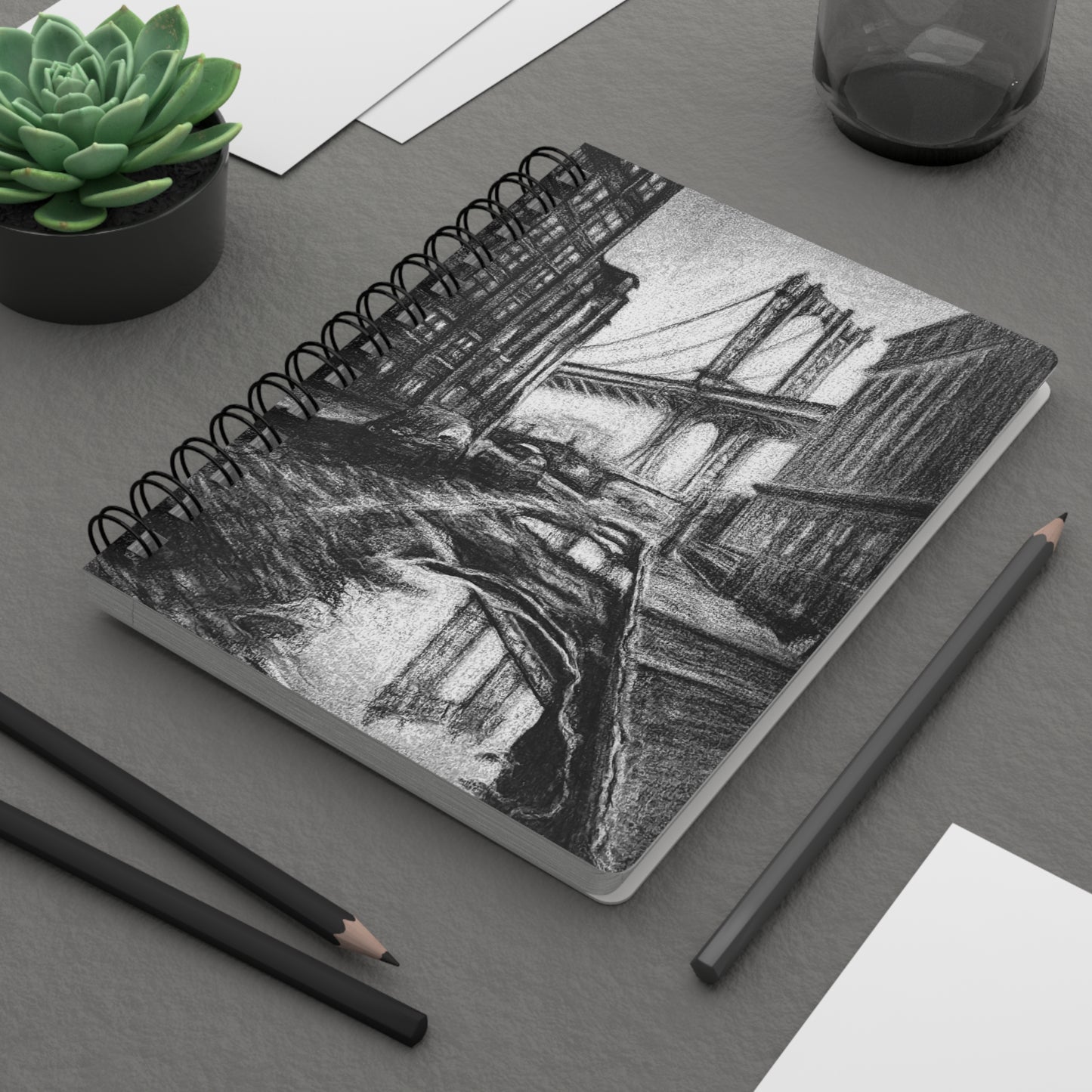 A spiral notebook with a charcoal drawing of an urban scene on the cover, featuring a bridge and buildings. On a desk.