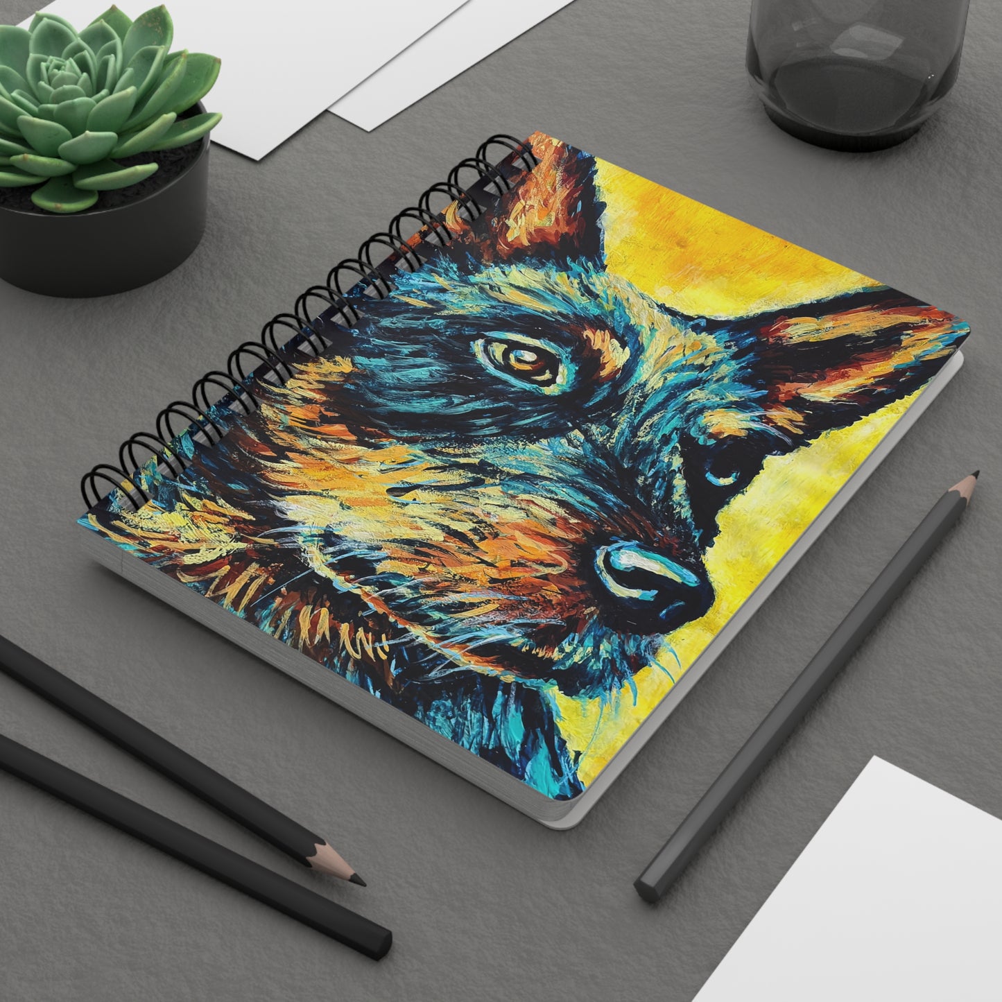 A spiral notebook with a colorful artistic portrait of a Blue Heeler dog on the cover. On a desk.