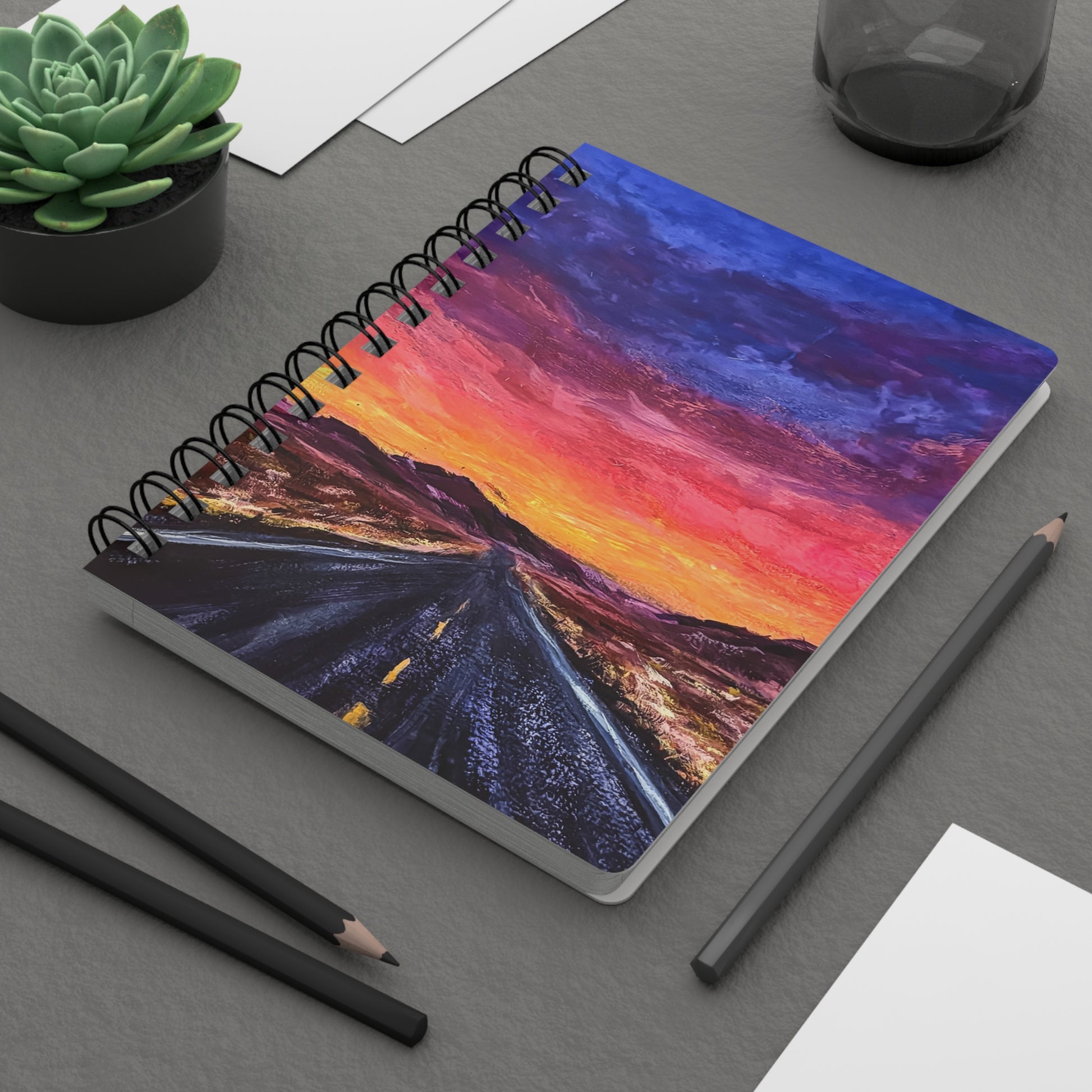 A spiral notebook with a cover depicting a fiery sunset over a desert landscape with a road stretching into the distance. On a desk.