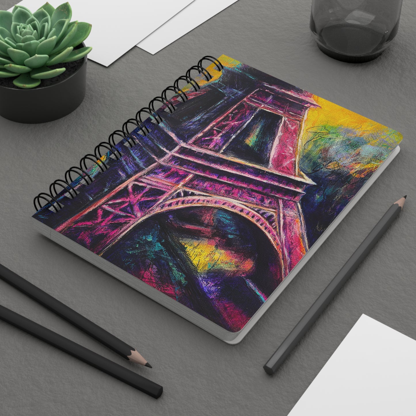 A spiral notebook with a colorful artistic cover featuring the Eiffel Tower with vibrant pinks and yellows and purples on desk
