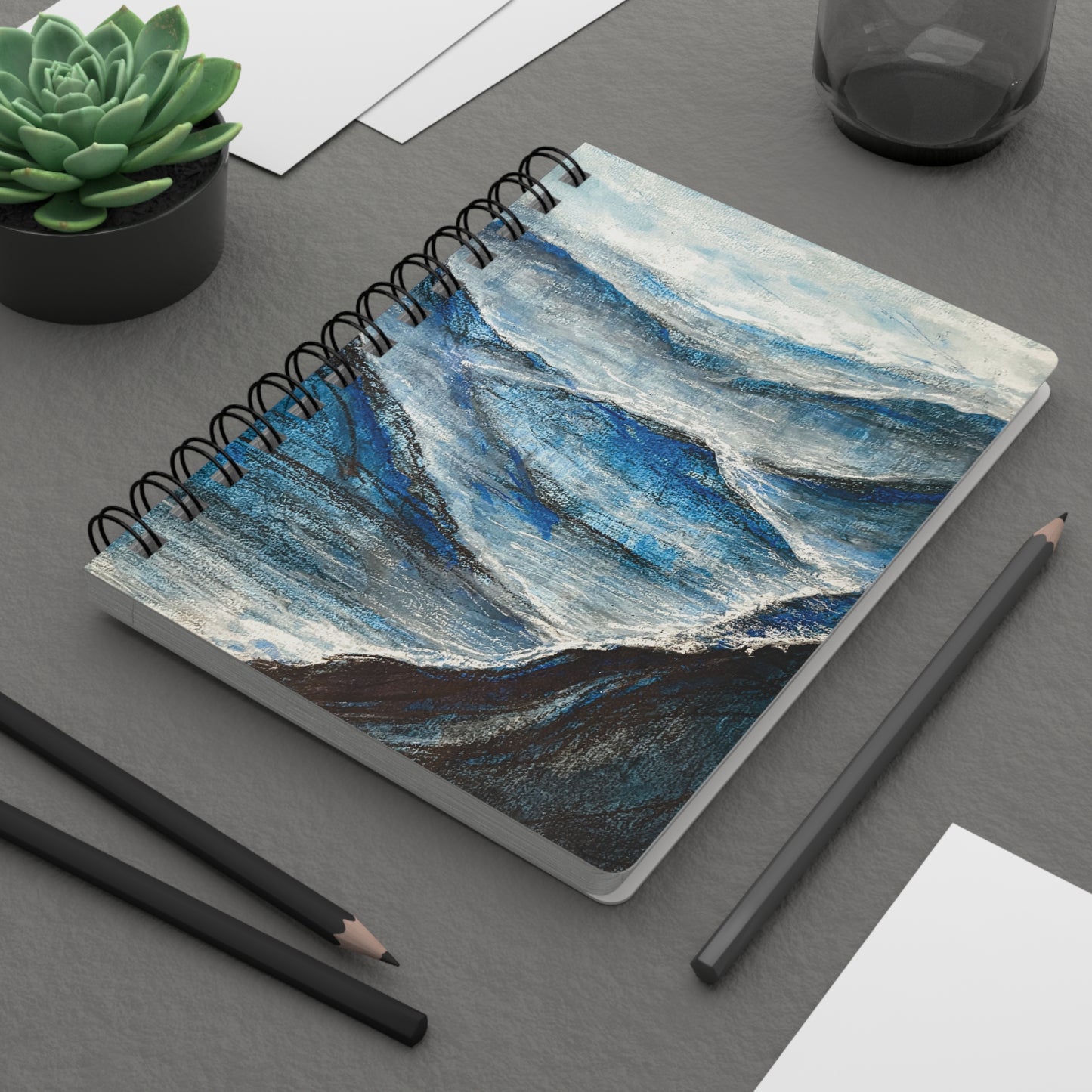 A spiral notebook with a abstract charcoal drawing with ethereal white and blue gouache accents, depicting a misty mountainous landscape. On a desk.