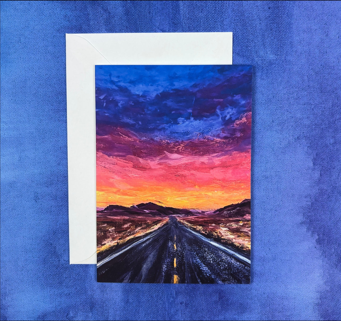 Greeting card with painting of a vibrant desert sunset over a road on a textured blue background