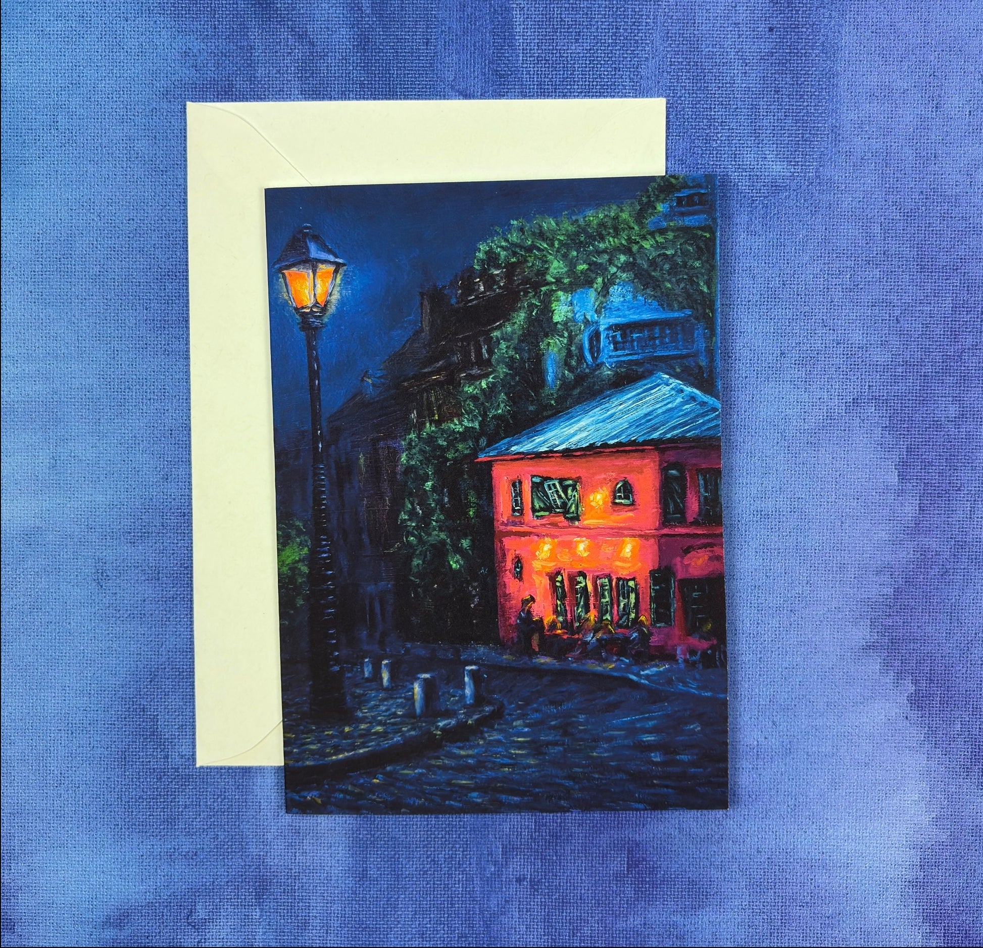 Greeting card with a painting of a pink cafe at night with cobblestone path on a blue textured background