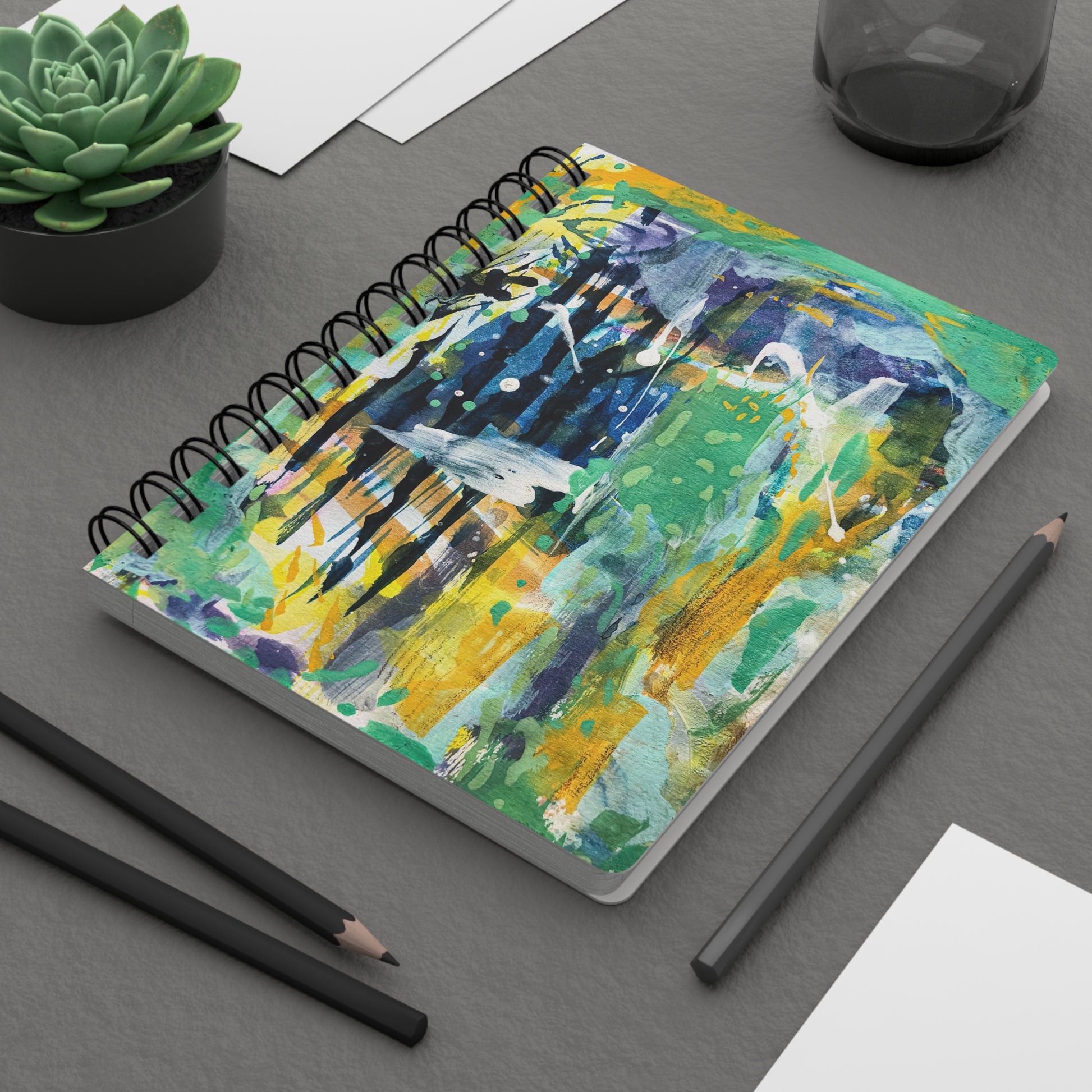 A spiral notebook with a colorful abstract pattern on the cover with green yellow blue and white. On a desk.